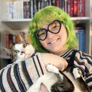 image of a nonbinary person (me) with green hair, heart-shaped glasses, and a striped sweater. they are hugging a startled looking white and brown tabby cat.
